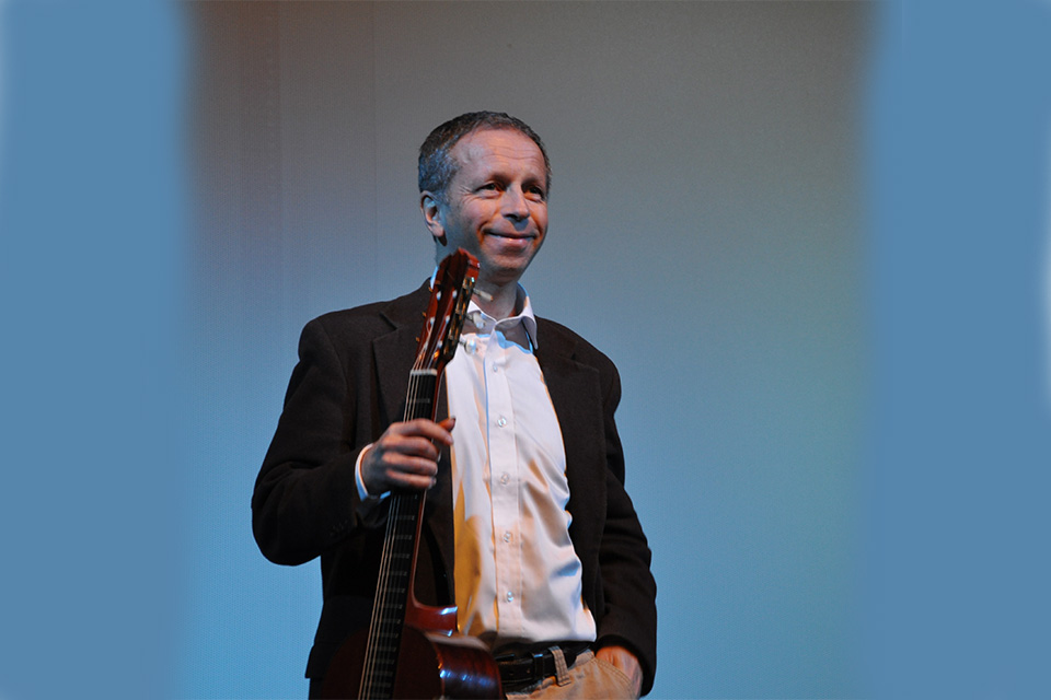 Goran Sollscher stands against a blue background holding a guitar