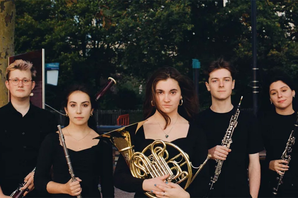 Members of the Aeolian Winds stand outside dressed in black and holding their instruments, from left to right: bassoon, flute, horn, oboe and clarinet