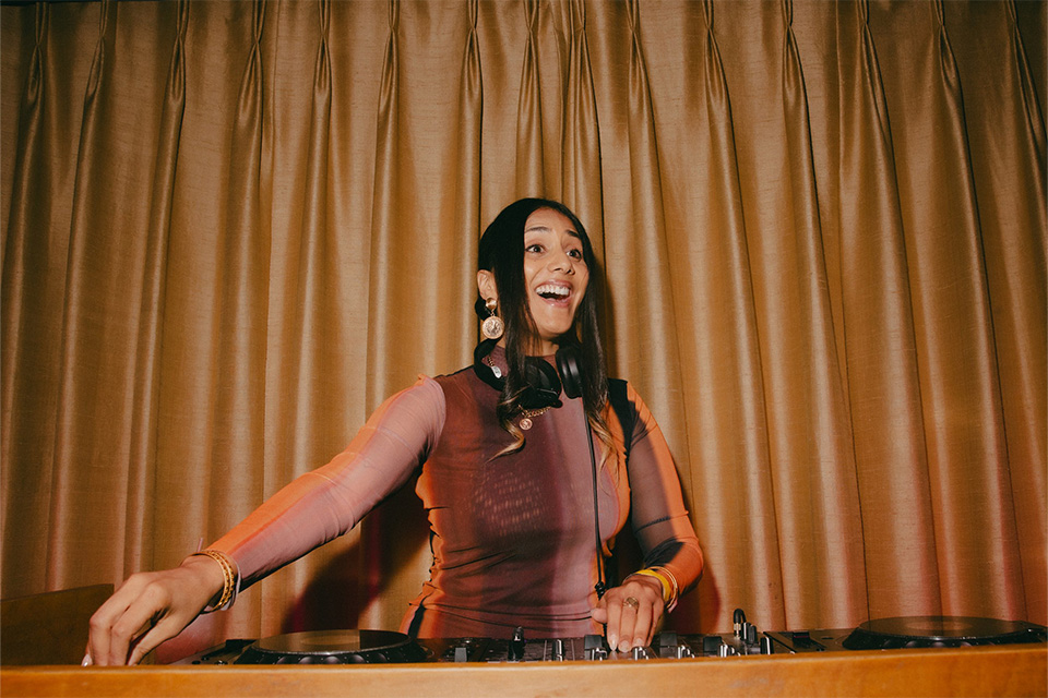 DJ Almass Badat performs in a DJ booth with headphones around her neck, smiling