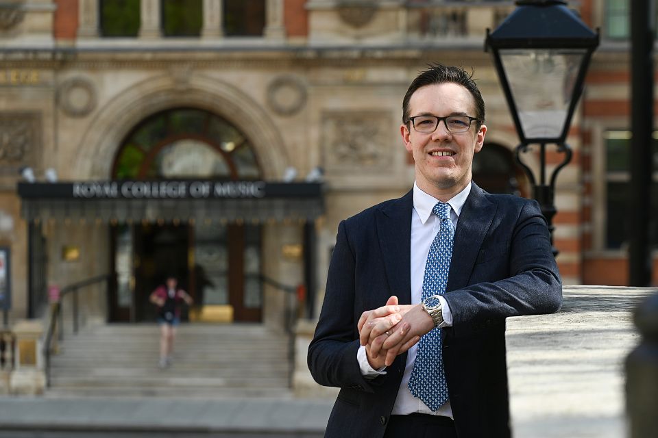 Andrew Moore wears a suit and leans on a wall in front of the Royal College of Music