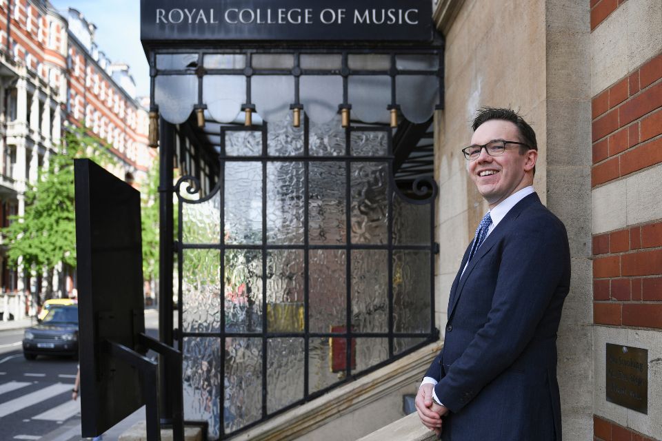 Andrew Moore wears a suit and stands on the steps of the Royal College of Music, smiling