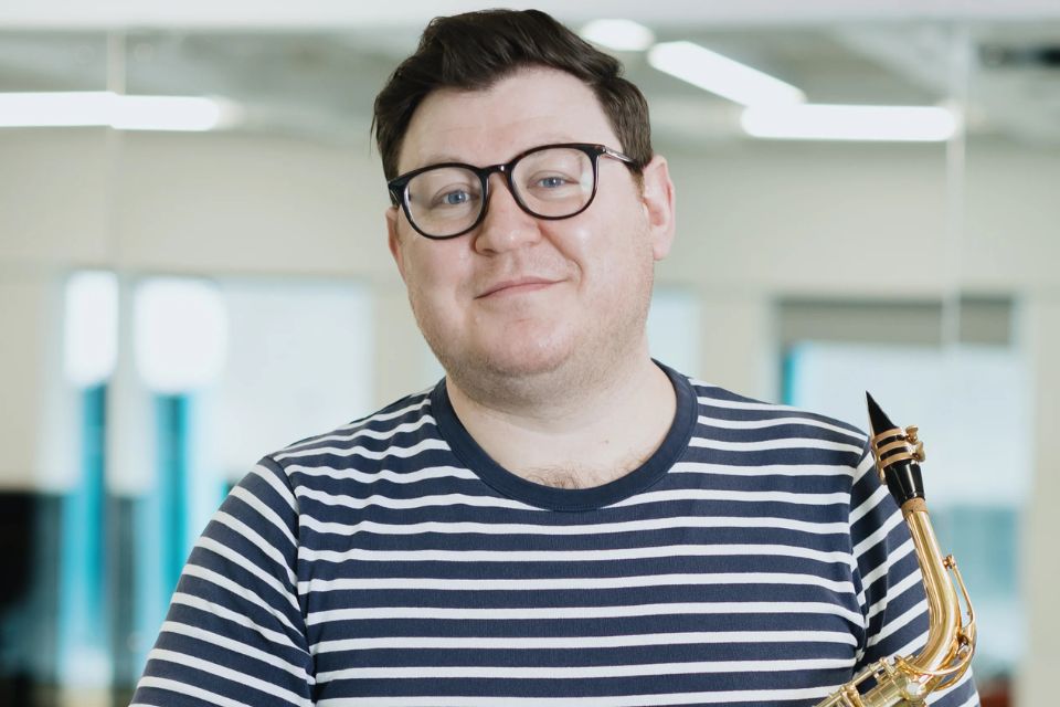 Andrew Somerville wears a black and white striped top and black glasses, holding his saxophone and smiling