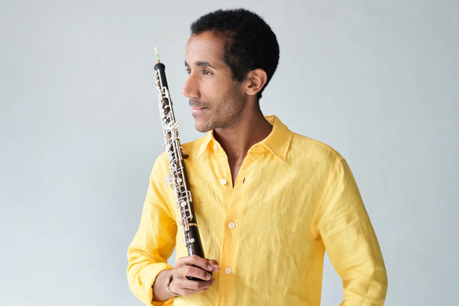 Armand Djikoloum sits against a dark orange wall in a suit, smiling at his oboe which is standing next to him