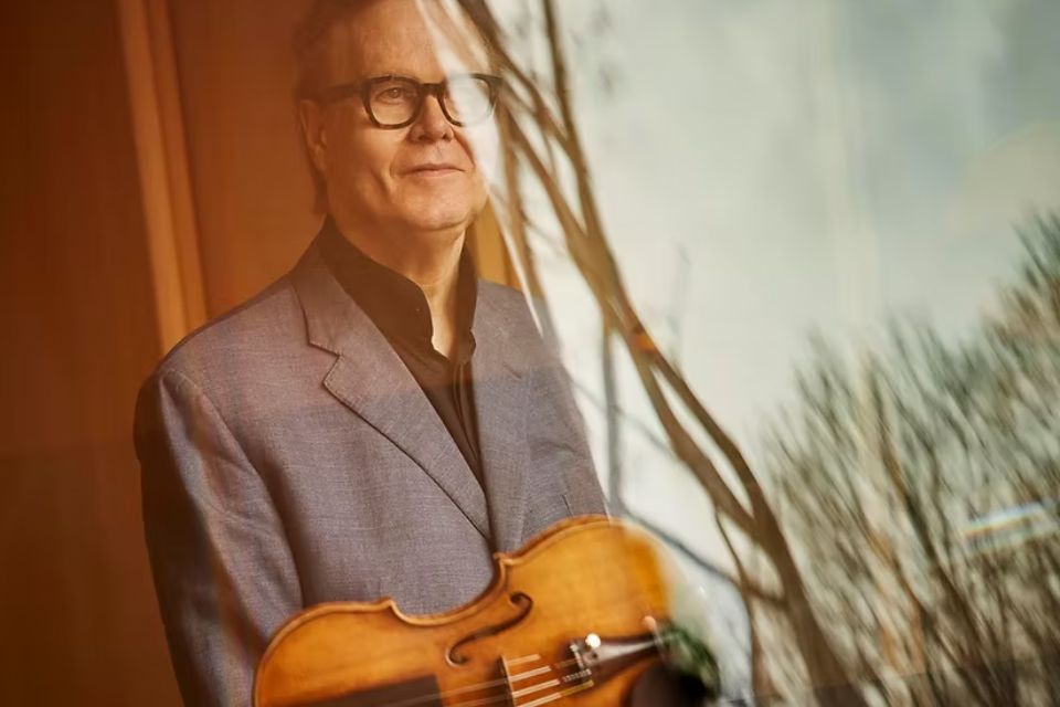 Augustin Dumay holds a violin and looks out a window, a reflection of trees in front of him