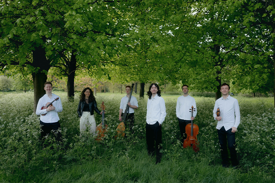 The six members of Bellot Ensemble wear white and stand in a green park holding their instruments