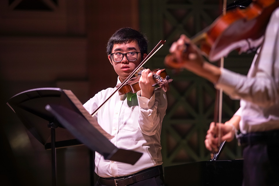 A boy plays his violin on a stage, looking over to another violinist next to him