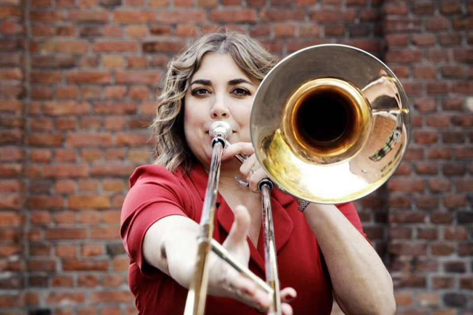 Clare Farr wears a red top against a brick backdrop and plays her trombone looking at the camera