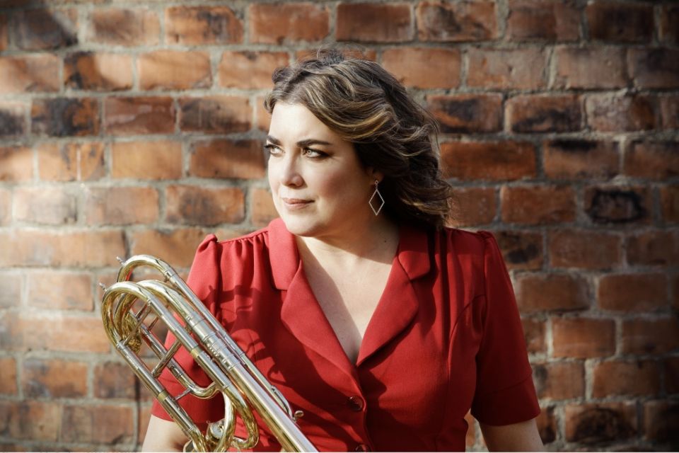 Clare Farr wears a red top against a brick backdrop and holds her trombone, looking to the side