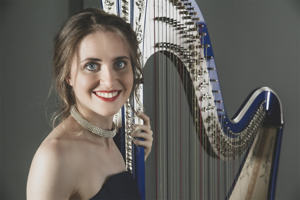 Claudia Lucia Lamanna wears a blue dress and red lipstick and holds her harp, smiling at the camera