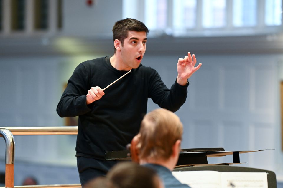 A conductor wears black and stands on a podium, gesturing passionately