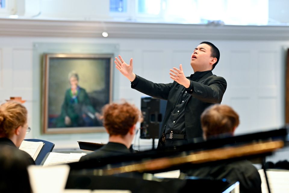 A conductor stands on stage wearing black with his hands in the air