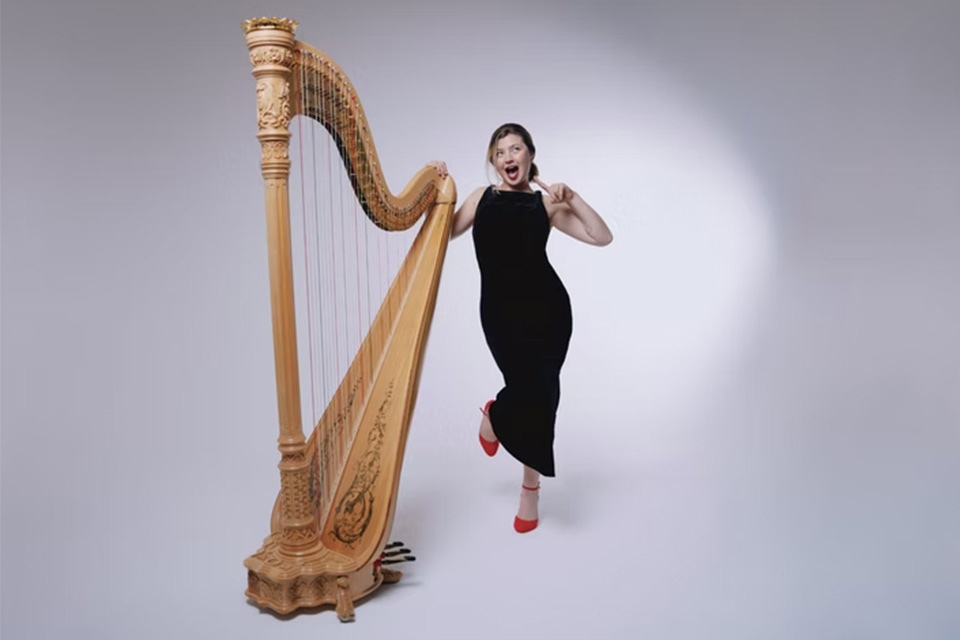 Emily Hoile stands next to her harp in front of a light grey backdrop