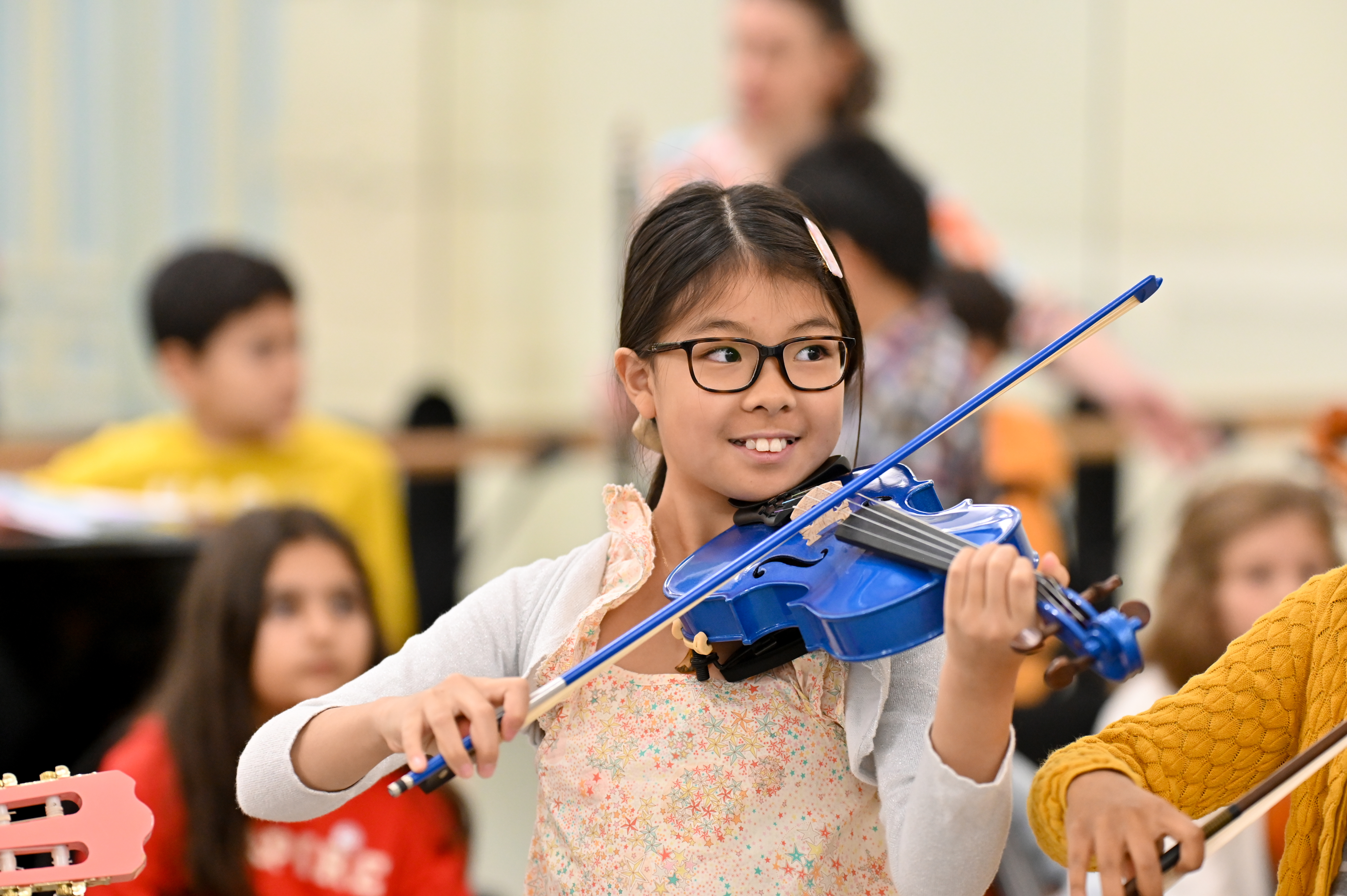 A little girl smiling and playing a bright blue violin with other children in the background