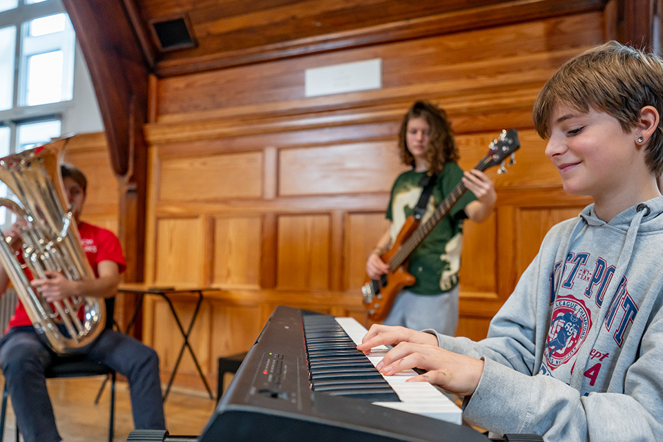 A young girl plays the keyboard and smiles, a tuba player and guitarist playing in the background