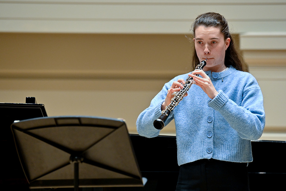 An oboist wears a blue cardigan and performs her instrument on stage