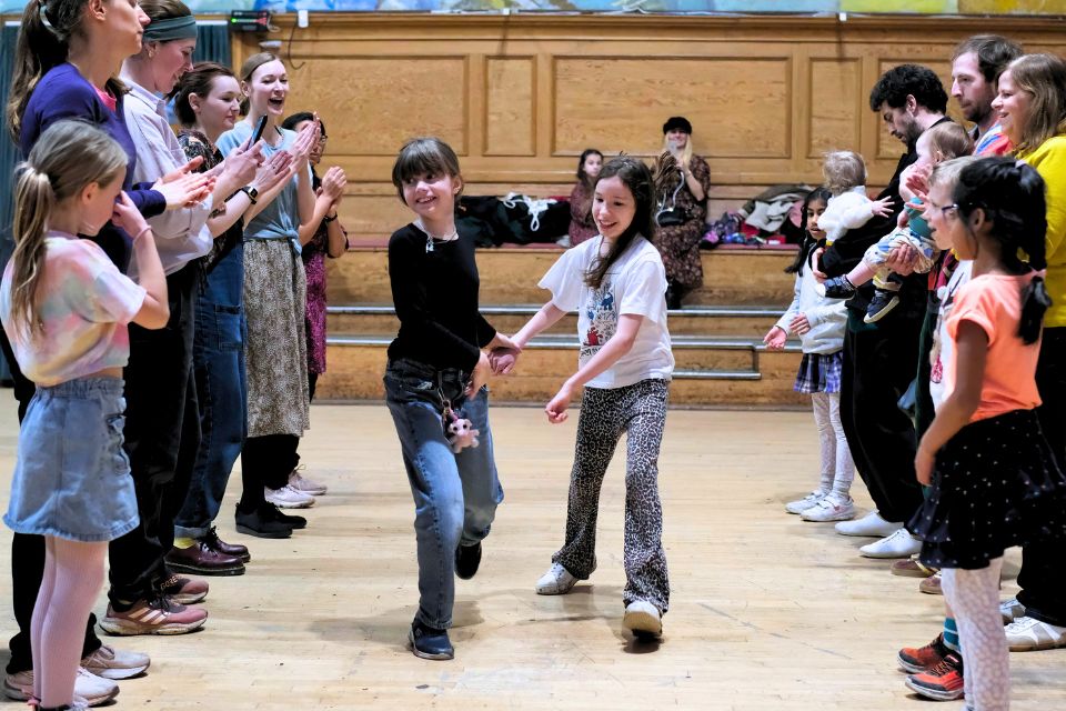 Two young girls dance holding hands while two rows of people clap either side