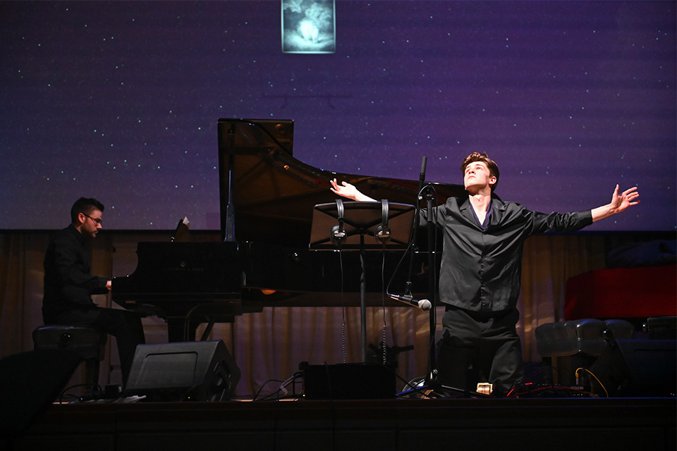 A man holds his arms out and looks up on a stage with a piano behind him