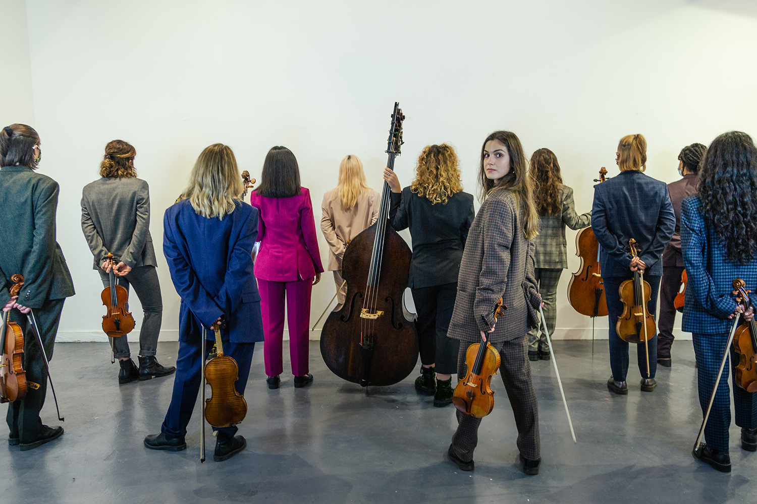 A group of people wearing suits, holding string instruments and standing with their backs to the camera with one woman turning around to look into the camera