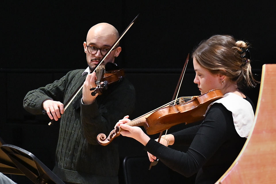 A young man and woman play baroque violins in a dark room
