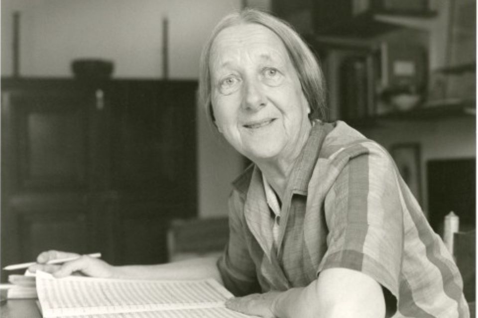 A black and white image of Imogen Holst smiling, a music score on the table in front of her