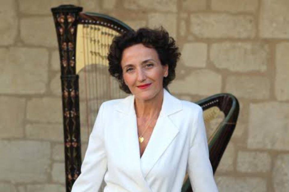 Isabelle Moretti wears a white top and stands in front of her harp against a light brick wall
