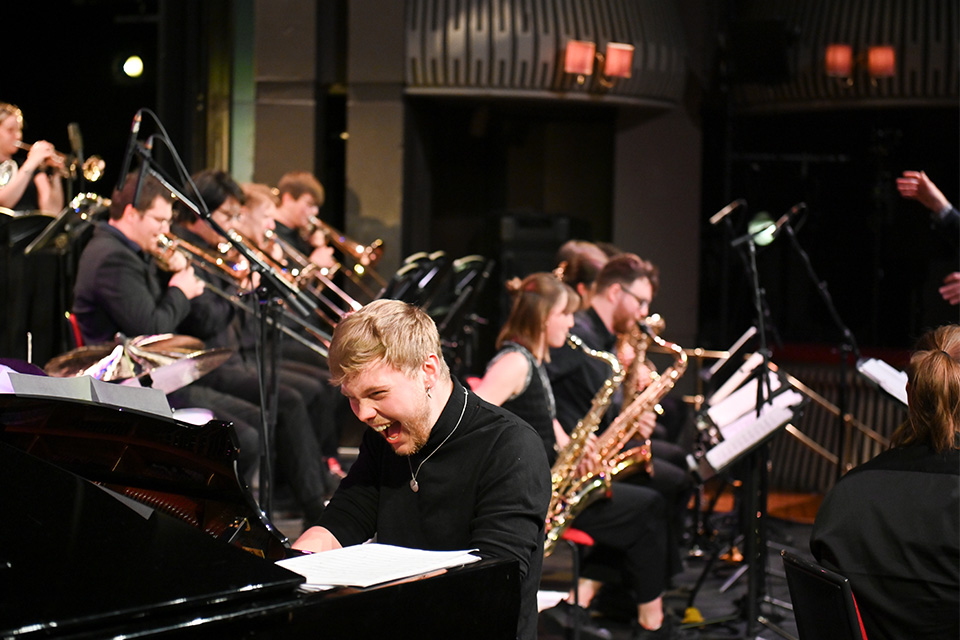A pianist performs on stage alongside a jazz orchestra, smiling excitedly