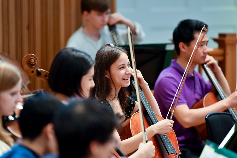 A young girl laughs as she plays the cello amongst other musicians in an orchestra