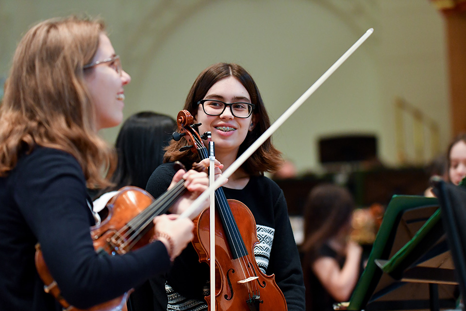 A young girl holds her violin and smiles at another violinist next to her