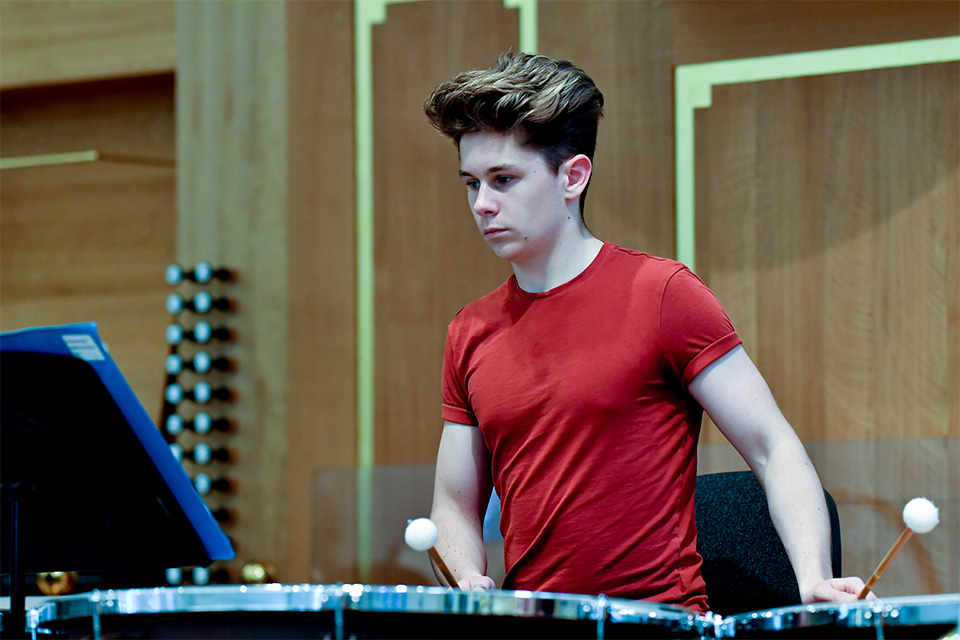 A young boy in a red t-shirt plays the timpani