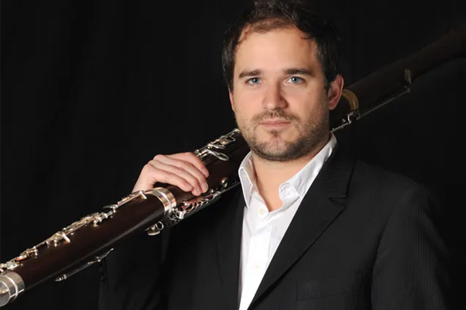Julien Hardy wears a dark blazer and white shirt and holds his bassoon against a dark background