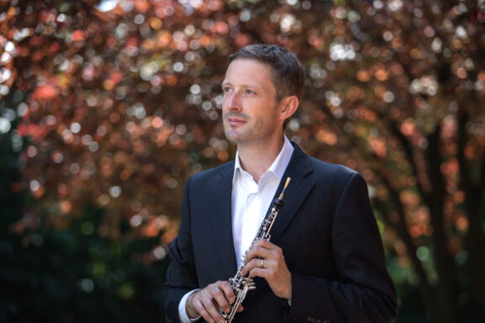 Kai Frömbgen stands outside in a shirt and blazer holding his oboe, against a backdrop of autumnal red leaves