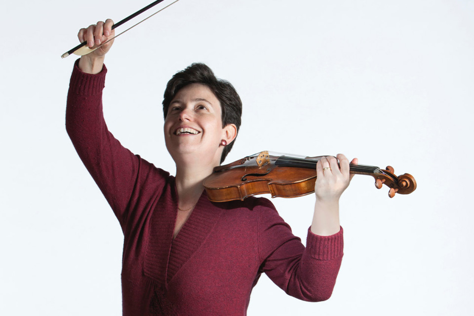 Kati Debretzeni wears a maroon shirt and holds her violin under her chin and her bow in the air, looking up and laughing