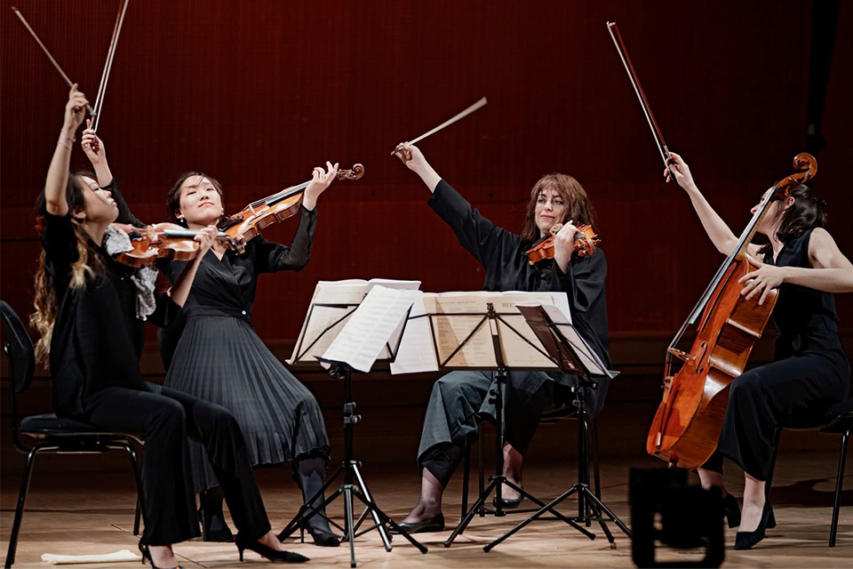 A string quartet performing on stage hold their instruments with their bows in the air