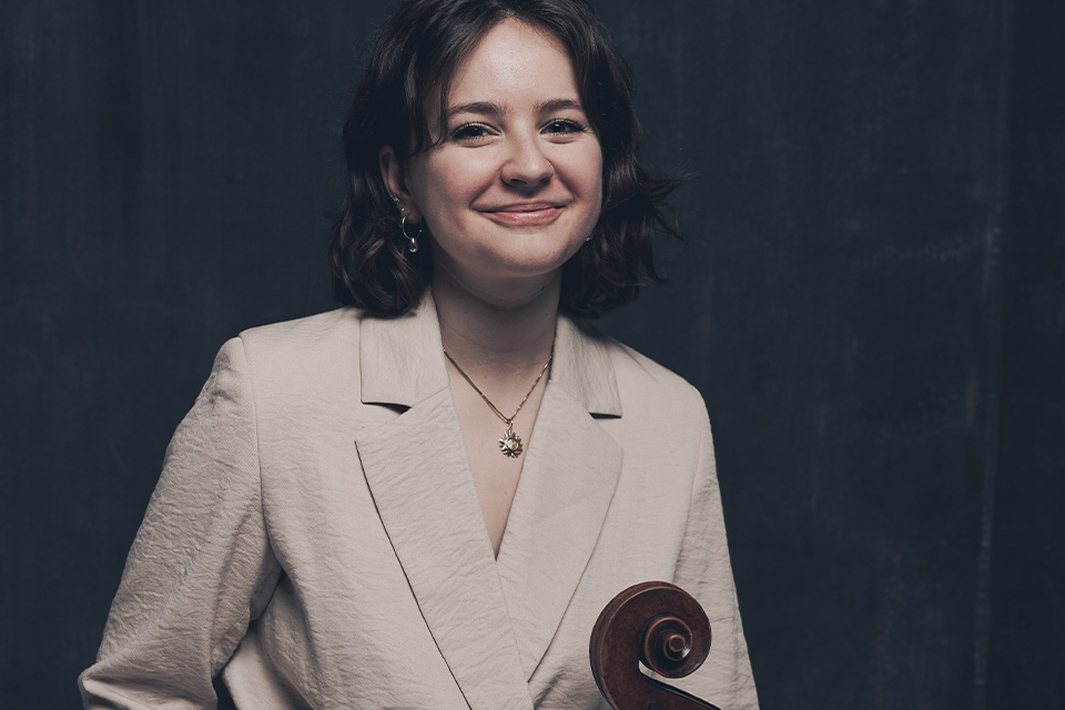 Laura can der Heijden wears a cream linen blazer and holds her cello, smiling