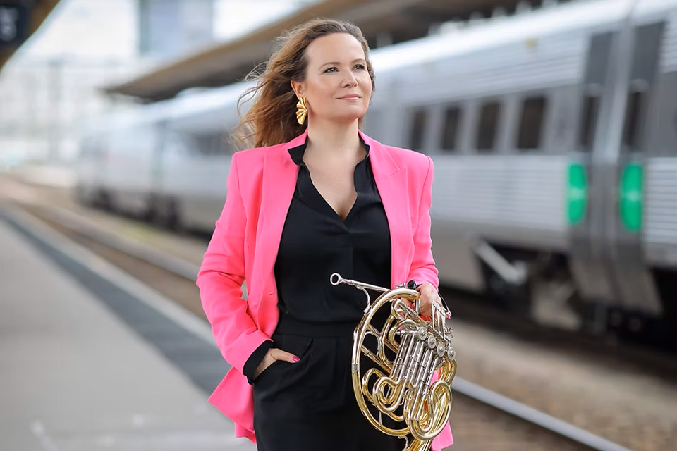 Lene Skomedal wears a pink blazer and stands on a train platform, holding her french horn