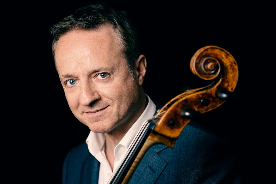 Marc Coppey wears a white shirt and blue jacket and holds his cello, smiling