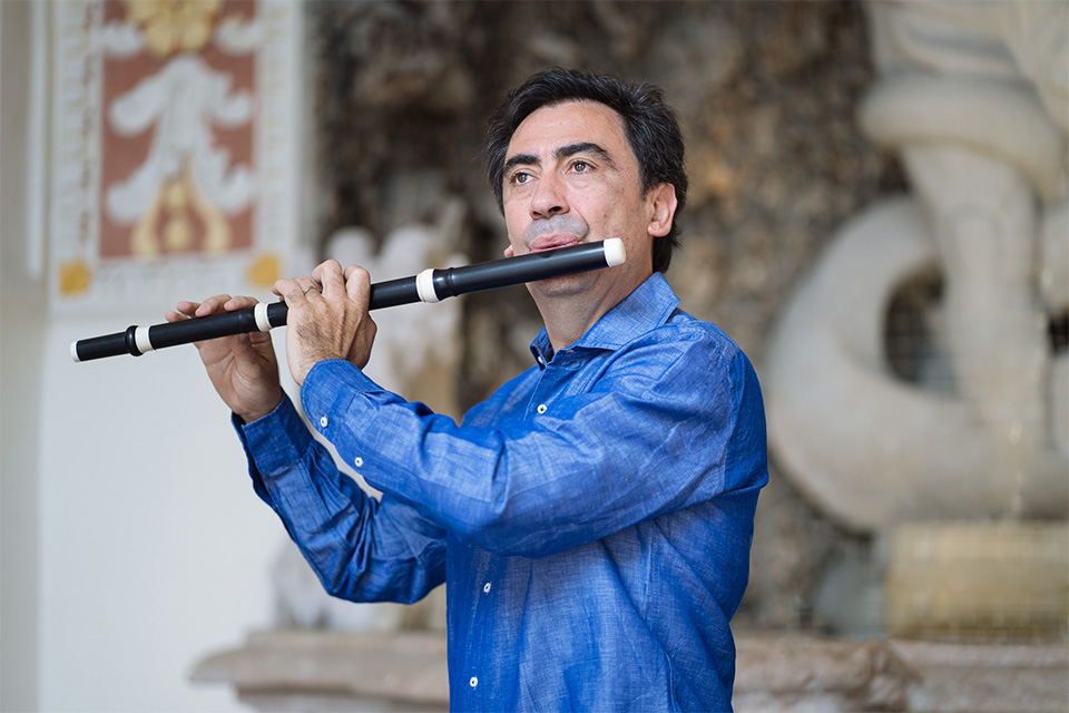 Marcello Gatti wears a bright blue shirt and plays his flute in an ornate room