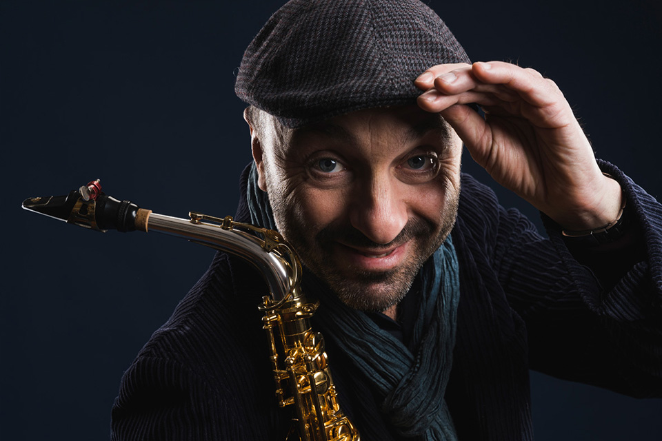 Marco Albonetti wears a flat cap and holds the edge, smiling, with his saxophone in the other hand