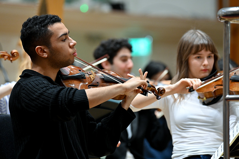 A young man and woman play violins as part of an orchestra on stage