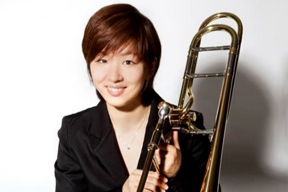 Mayumi Shimizu holds her trombone and smiles, wearing black against a white background