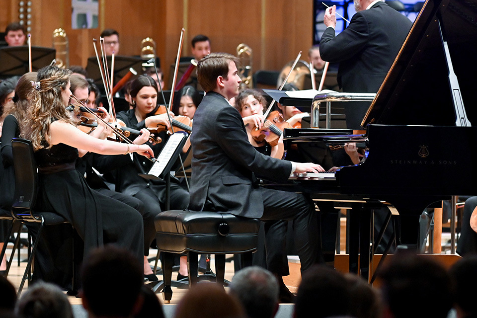 Misha Kaploukhii wears a suit and plays the piano in front of an orchestra on stage