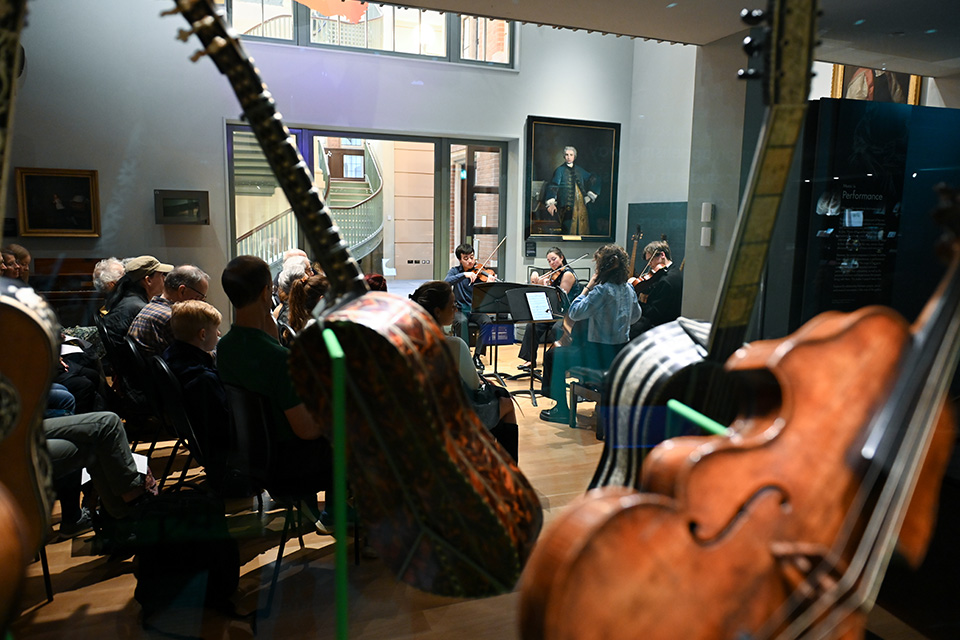 A string quartet performs to an audience in the RCM Museum, captured between exhibit displays of historical string instruments