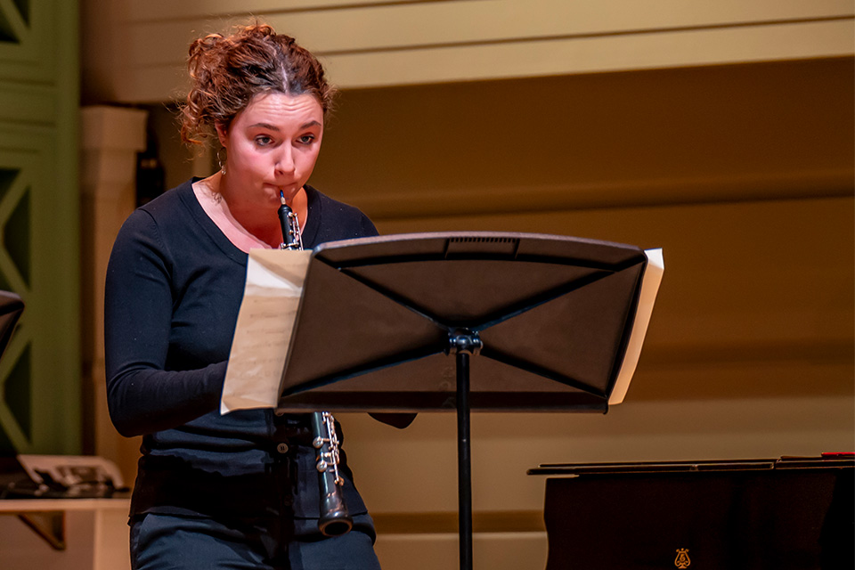 An oboist wears black and performs on stage in front of a music stand