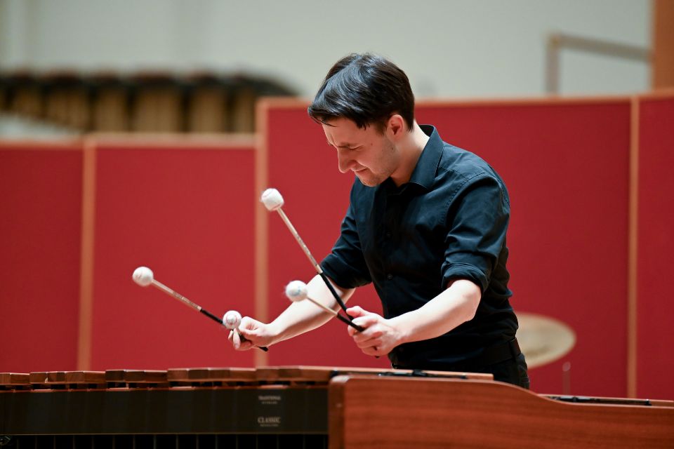 A young man wearing a black shirt holds four mallets and plays the glockenspiel