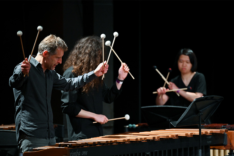 Three percussionists play the glockenspeil wearing all black, their mallets rasied in the air