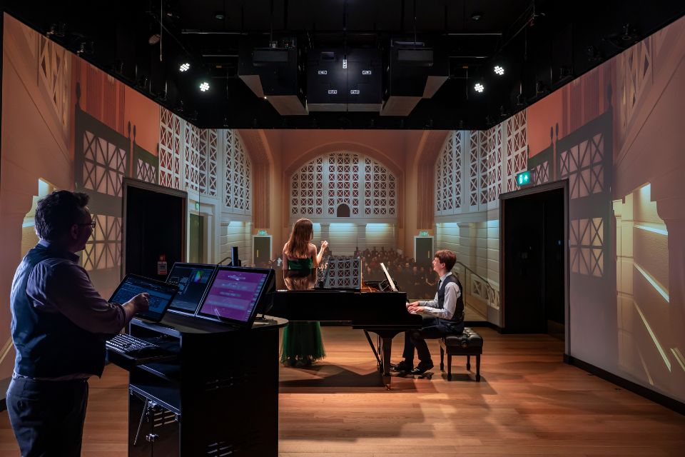 A pianist and vocalist perform in front of a virtual audience projected on a screen