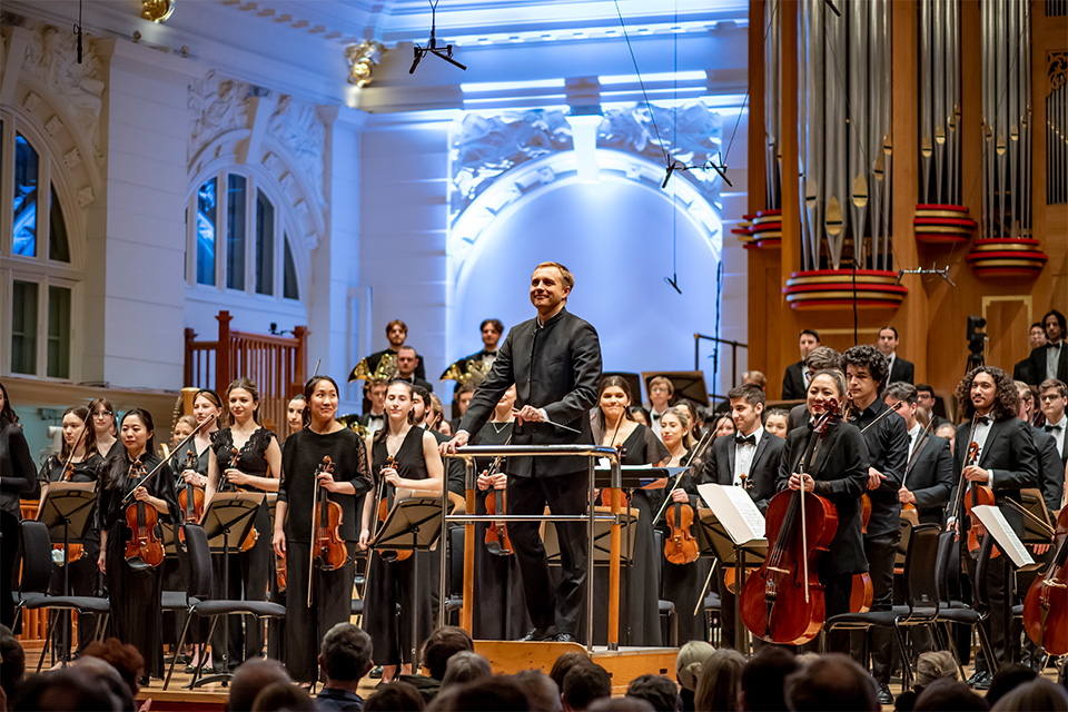 Vasily Petrenko stands on the conductors podium and looks out to the audience, the orchestra also standing behind him