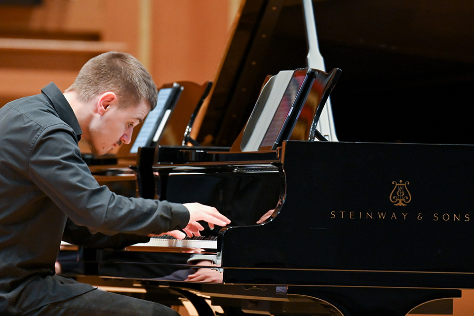 A young man wearing a black shirt plays the piano passionately on stage