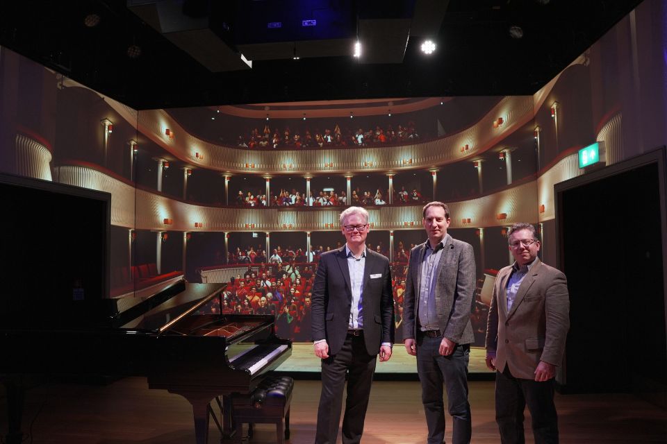 Three men stand next to a grand piano and in front of the RCM's Performance Simulator screen