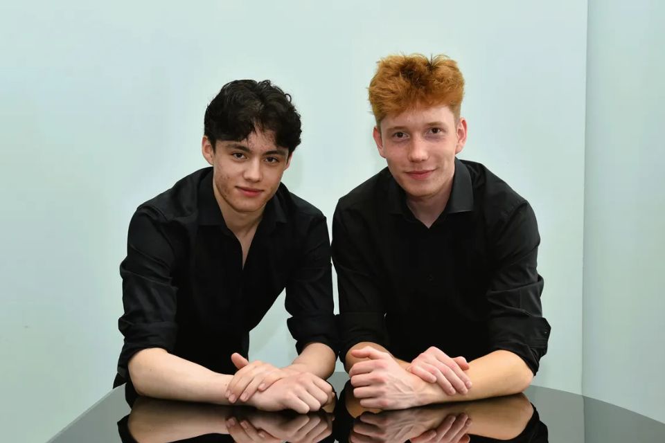 Two boys wear black and lean on a piano against a turquoise background
